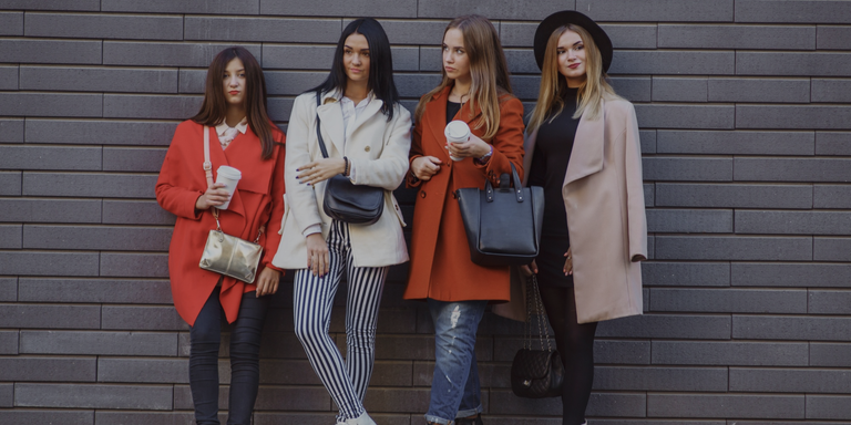 Four women standing against a brick wall, each holding a different colored handbag.